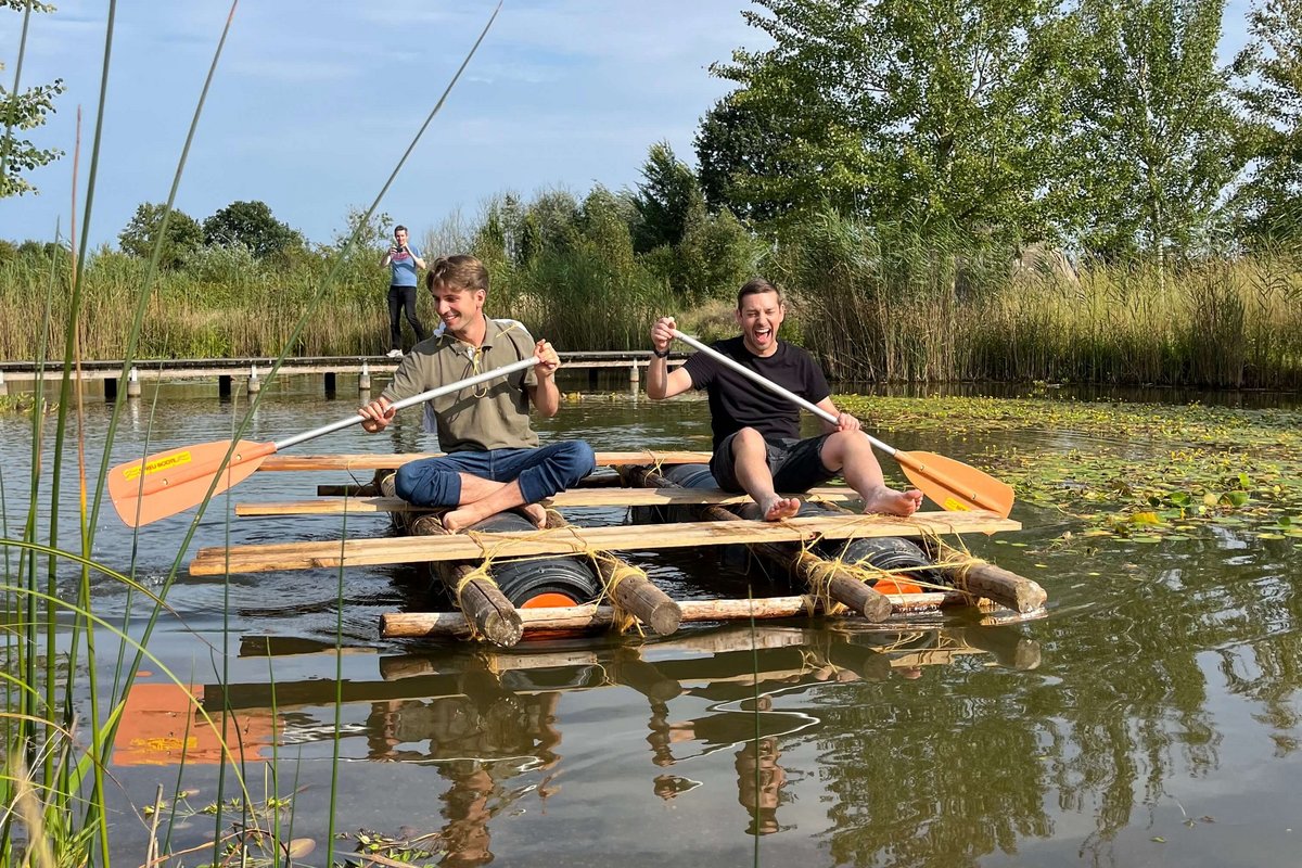 Zwei Personen sitzen auf einem selbstgebauten Floß und paddeln auf dem Teich im Garten der Stille. 